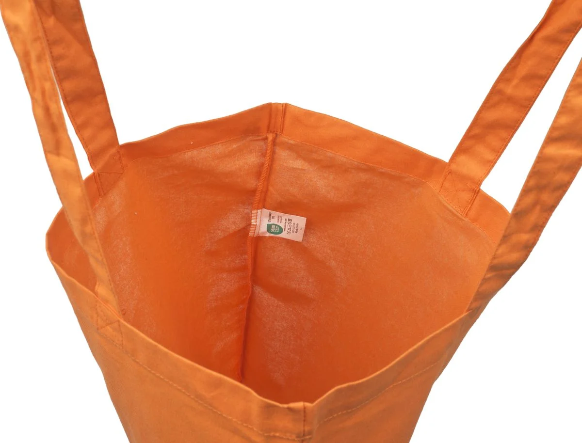 Oranžinio 140 g/m² medžiaginio maišelio iš medvilnės vidaus vaizdas, kuriame matyti audinys be pamušalo, susiūtos siūlės ir viduje pritvirtinta maža etiketė.